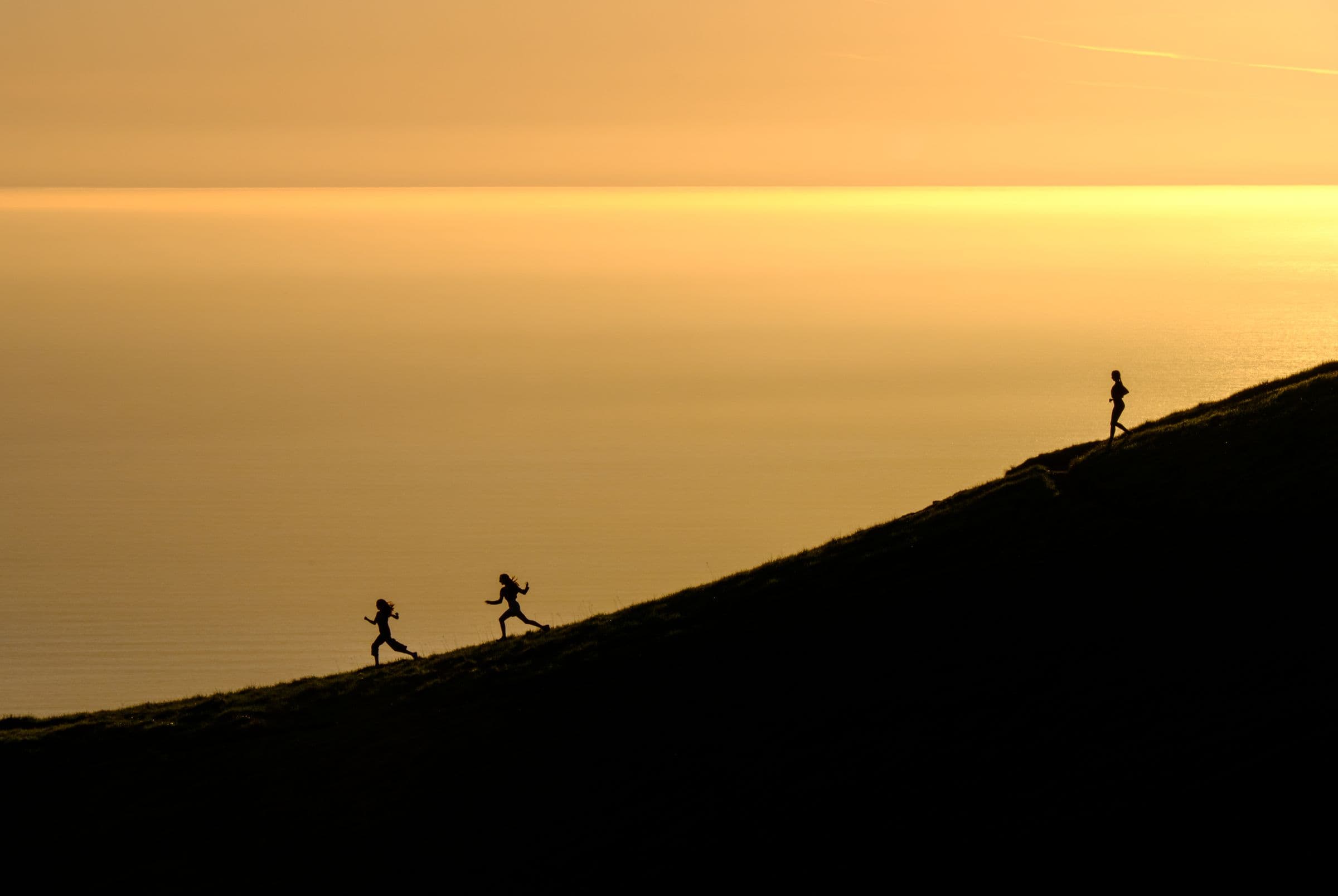 Três corredores em silhueta numa encosta ao pôr do sol, com o mar dourado ao fundo